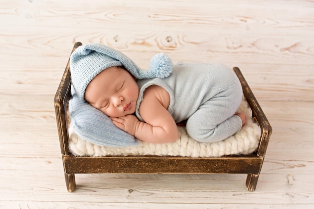 A cute newborn boy in the first days of life sleeps in a blue overalls on a wooden brown bed Blue knitted hat and Blue head pillow Against a light wooden background High quality photo