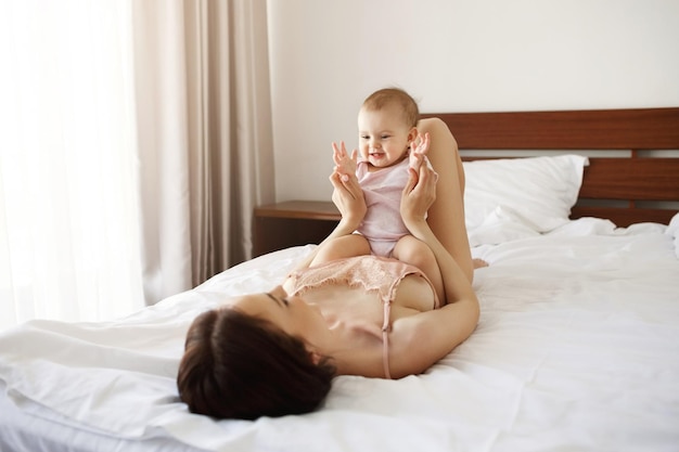 Cute nice baby daughter sitting on her young mom lying on bed smiling at home