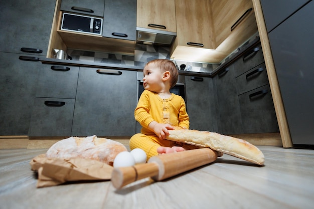 A cute oneyearold boy is sitting in the kitchen and eating a long bread or baguette in the kitchen The first eating of bread by a child Bread is good for children