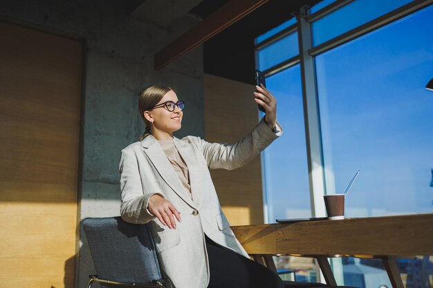 Cute positive female employee with long blond hair in casual clothes looking at phone while working on new business project at table with laptop and gadgets in office