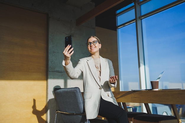 Cute positive female employee with long blond hair in casual clothes looking at phone while working on new business project at table with laptop and gadgets in office