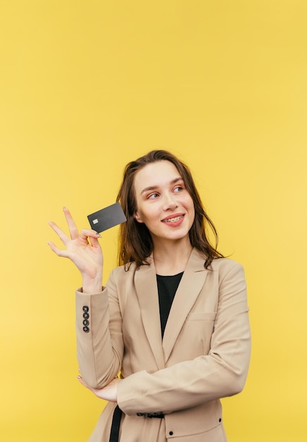 Cute positive lady in a beige jacket poses with a bank card on a yellow background and looks away