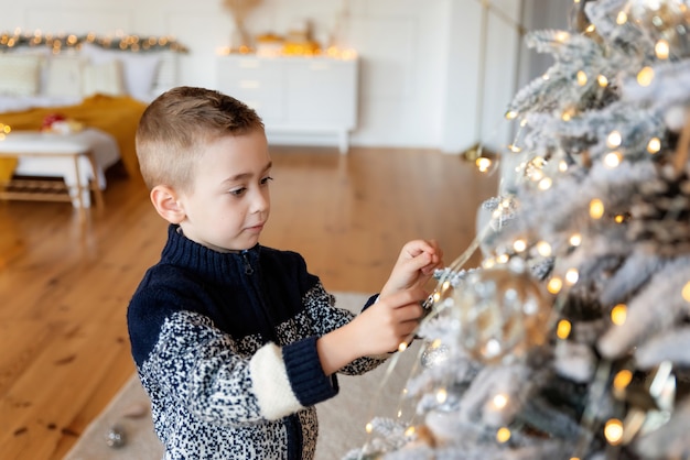 Cute prechooler Caucasian boy child decorating Christmas tree in room. Xmas party celebration. Concept of New Year atmosphere and holiday mood.