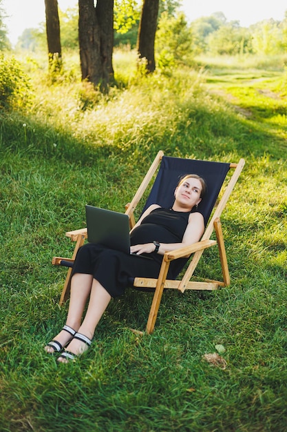 A cute pregnant woman is sitting in a park and working remotely on a laptop Online work on maternity leave Modern pregnant woman