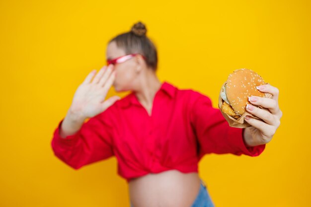 Photo a cute pregnant woman in a pink shirt and pink glasses eats fast food a pregnant woman on a yellow background with an appetizing burger in her hands harmful food for pregnant women