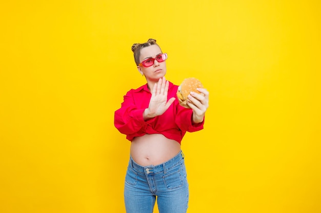 Photo a cute pregnant woman in a pink shirt and pink glasses eats fast food a pregnant woman on a yellow background with an appetizing burger in her hands harmful food for pregnant women