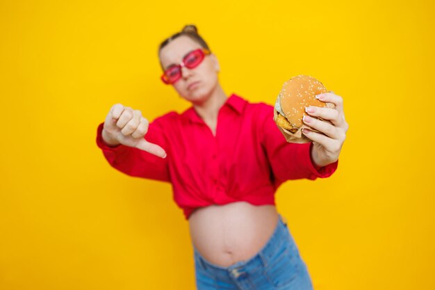 Photo a cute pregnant woman in a pink shirt and pink glasses eats fast food a pregnant woman on a yellow background with an appetizing burger in her hands harmful food for pregnant women