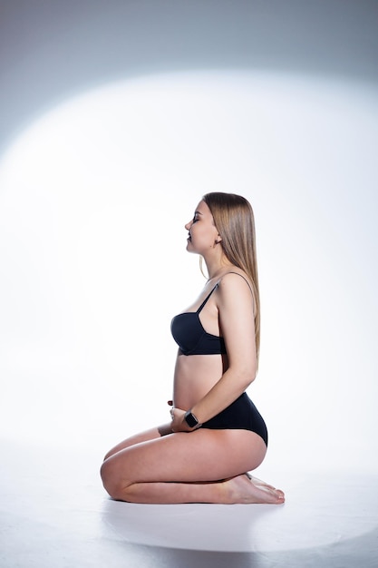 Cute pregnant woman with long hair in black underwear is sitting on a white background Women's underwear for pregnant women