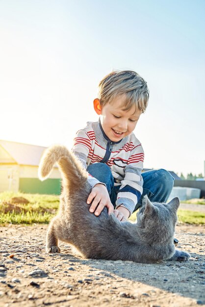 Cute preschooler boy with blond hair plays with grey domestic cat in country yard