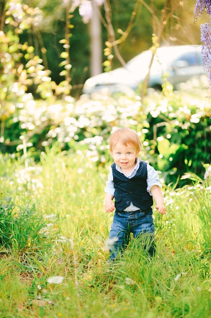 Cute red-haired baby boy is standing on a green grass in the park on a sunny day