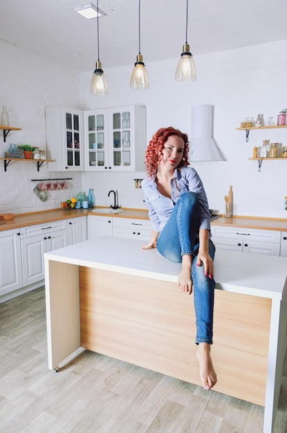 Photo cute redhead curly woman 30 in blue jeans and a shirt sitting, relaxing in bright kitchen. housewife