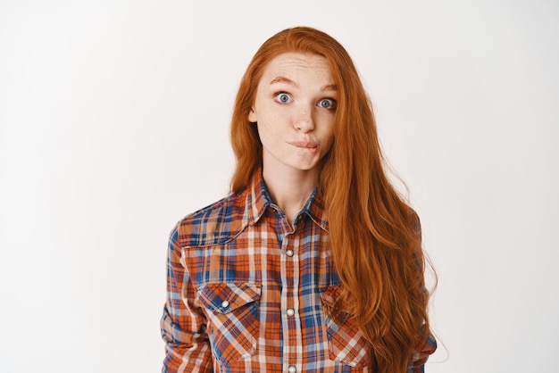 Cute redhead girl standing indecisive over white background sucking lips and staring at camera surprised