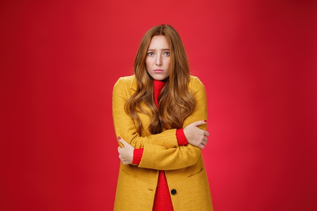 Cute and sad trembling redhead girl in light yellow coat shaking from cold, having goosebumps frowning and pouting from sorrow as freezing on windy weather over red background. Copy space