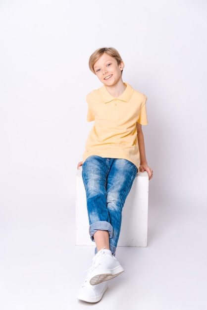 Cute schoolboy sitting on box wearing jeans and yellow tshirt isolated