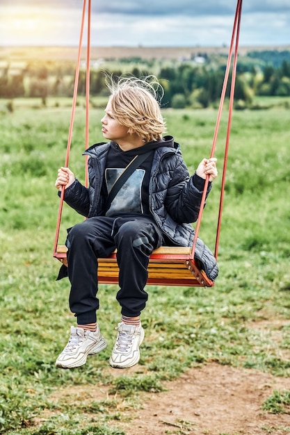 Cute schoolboy with long blond hair rides on swing looking aside on cloudy day in countryside