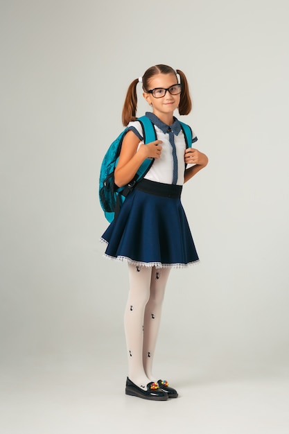 Cute schoolgirl in glasses with blue backpack looking at camera on grey background