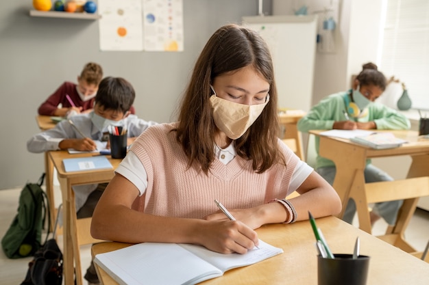 Cute schoolgirl in protective mask making notes