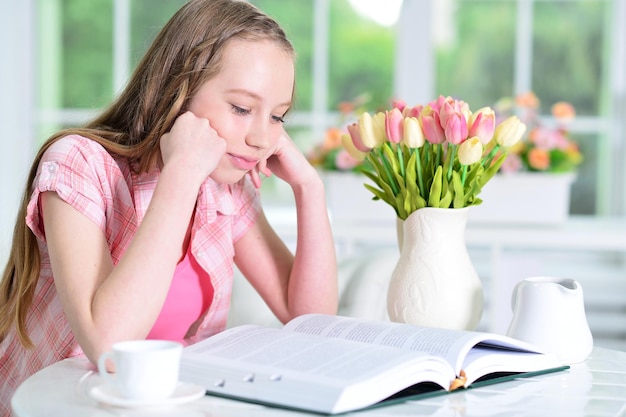 Cute schoolgirl sitting at table and reading at home