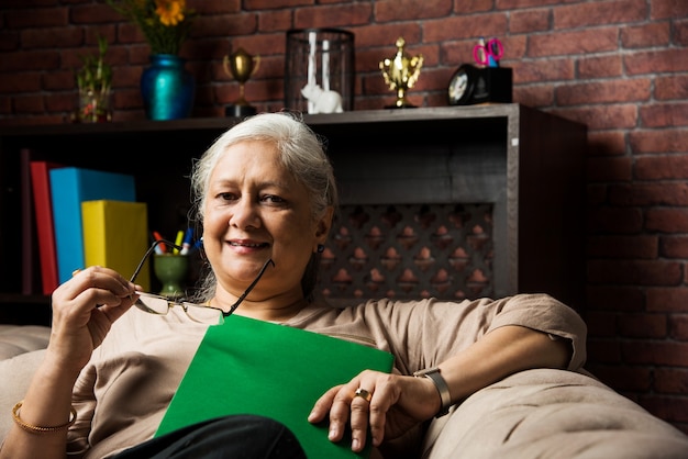 Cute Senior indian asian lady sitting on recliners chair or sofa reading book or using tab or laptop computer