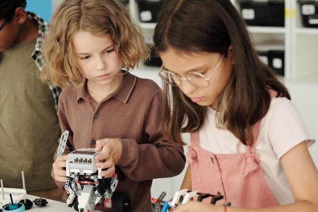 Cute sesrious schoolboy looking at his classmate constructing robot at lesson