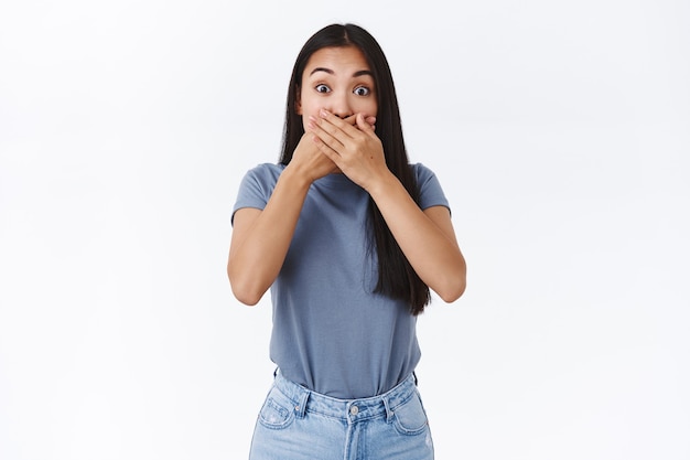 Cute silly amused asian girl heart awesome rumour, slap her mouth with hands and stare camera astonished, trying hold herself not tell anyone gossip, standing white wall surprised