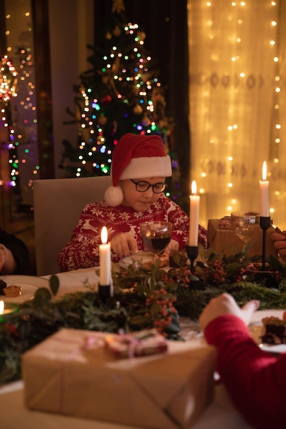 Cute small boy in Santa hat having his Christmas dinner in the evening at home near Christmas tree.