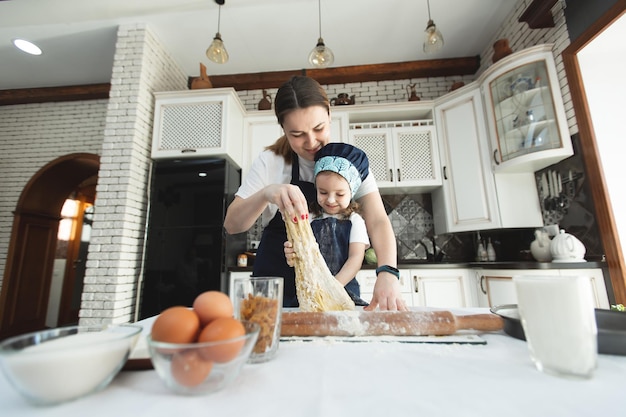 Cute small child daughter helping mum kneading preparing dough in bowl together in modern kitchen