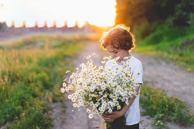 Cute smiling boy at camomile field at sunset in soft sunlight Life without allergies breathe freely Boy and daisies Child dreaming and smiling against the background of a camomile field