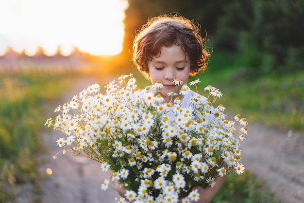 Cute smiling boy at camomile field at sunset in soft sunlight Life without allergies breathe freely Boy and daisies Child dreaming and smiling against the background of a camomile field