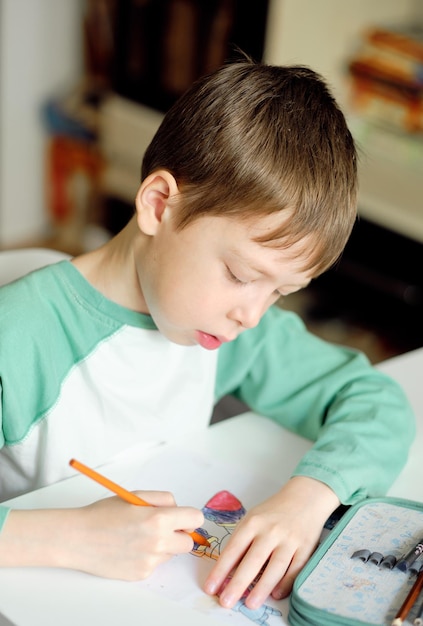 Cute and smiling cucasian boy draws with colored pencils while sitting at the table the boy paints the drawing with a blue pencil Drawing lessons Art therapy for stress relief