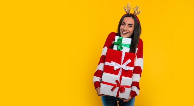 Cute smiling excited young brunette woman in deer hat, with many colorful Christmas gift boxes in hands isolated on yellow background.