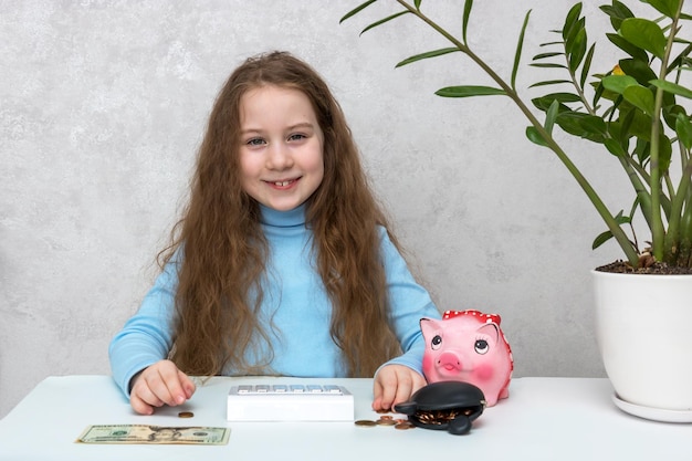 Cute smiling girl sitting at the table counts money using a calculator from a piggy bank and a wallet financial literacy