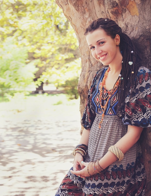 Cute smiling hippie indie style woman with dreadlocks,  dressed in boho style ornamental dress posing outdoor