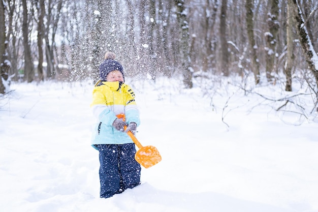 Cute smiling little boy with shovel playing with snow outdoors in winter forest