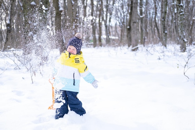 Cute smiling little boy with shovel playing with snow outdoors in winter forest