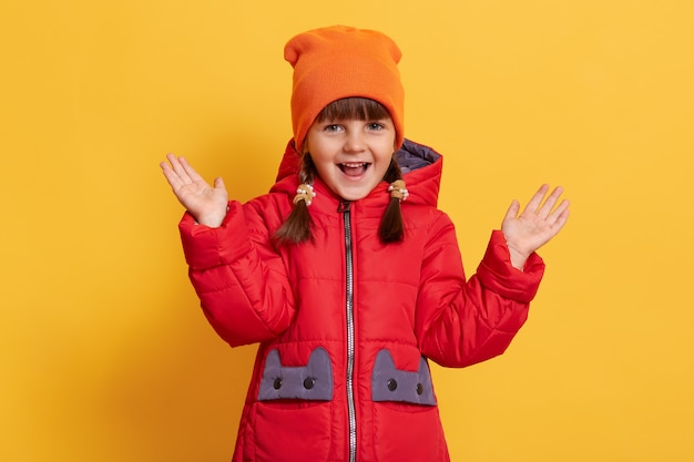 Cute smiling little child raising her hands up isolated on yellow wall, looks at front with opened mouthcap.