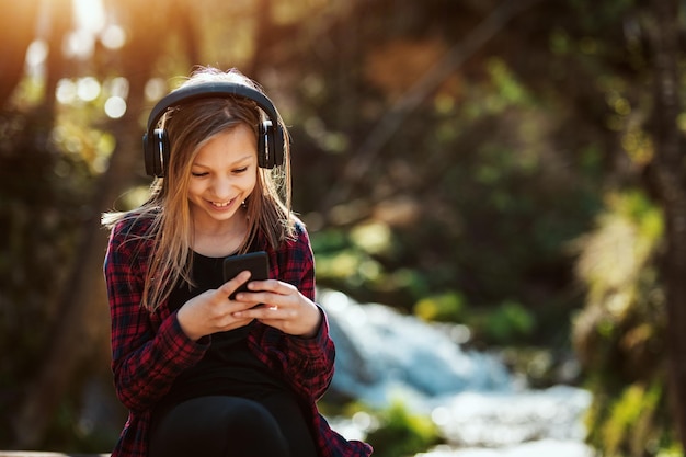 Cute smiling little girl listening music from a her smartphone and enjoying landscape at the beautiful waterfall in mountains.