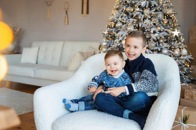 Cute smiling siblings children in sweaters and jeans hug near decorated Christmas tree at home. Two little happy kids, preschool boy and toddler baby. Brother love. Concept friendship.