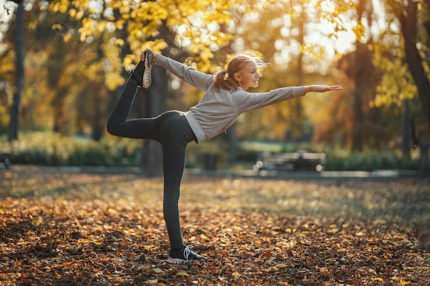 Cute smiling teenage girl is doing stretching exercise in the city park, and enjoying sunny autumn day.