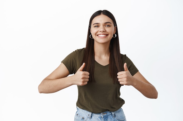 Cute smiling woman being supportive, showing thumbs up and looking happy, approve good choice, praise you, like or agree, standing over white wall