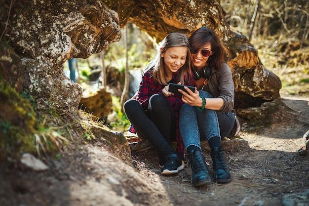 Cute smiling young woman and her little daughter with smartphone in their hands are looking at the smartphone and are enjoying landscape in mountains.