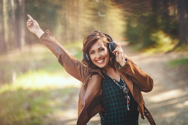 Cute smiling young woman is walking along forest path in early spring sunny day, having fun and listening music.