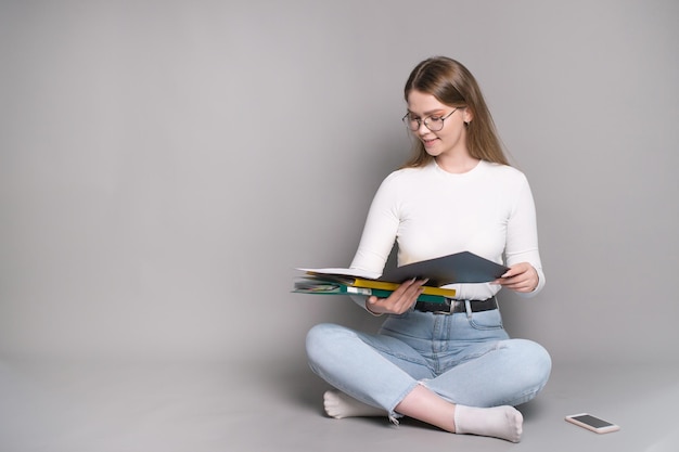 A cute student with glasses is sitting on a gray background and looking at folders for documents