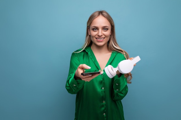 Cute stylish attractive blonde girl with headphones and smartphone stretches out her hands on a blue