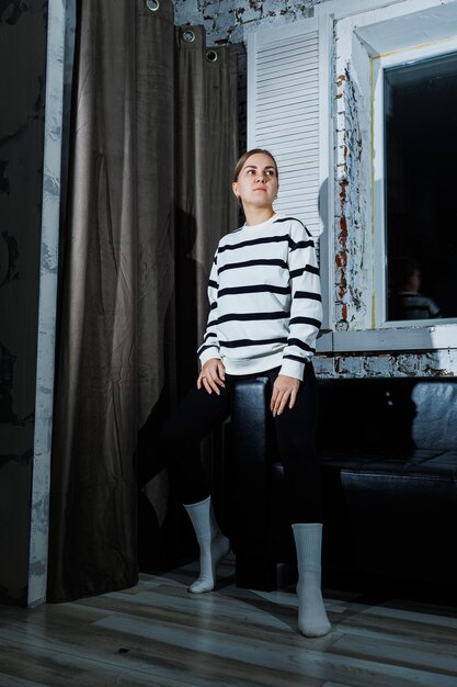 Cute stylish young woman in black leggings and striped jumper sitting on sofa Women's fashion women's casual style