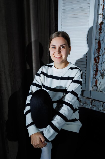 Cute stylish young woman in black leggings and striped jumper sitting on sofa Women's fashion women's casual style