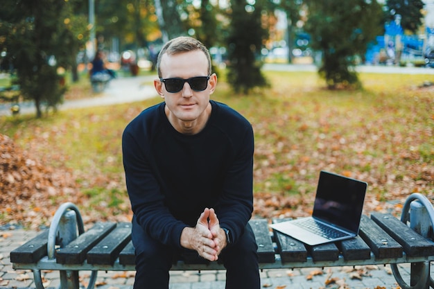 Cute successful young man in sunglasses sitting on park bench with laptop and phone and looking away on sunny day Freelance remote work concept