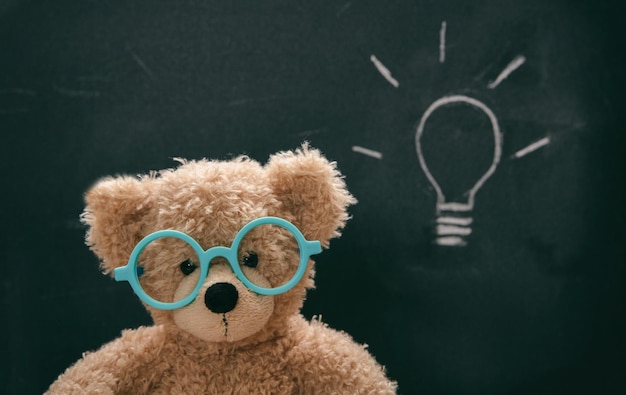 Cute teddy wearing glasses against blackboard with a lightbulb