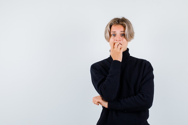 Cute teen boy in black turtleneck sweater biting nails emotionally and looking frightened , front view.