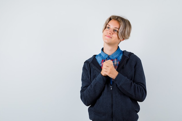 Cute teen boy clasping hands in praying gesture in shirt, hoodie and looking hopeful , front view.